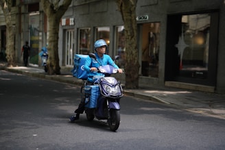 A delivery person wearing a blue uniform and helmet rides a scooter with large blue delivery boxes on a quiet street lined with trees and shops.