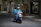 A delivery person wearing a blue uniform and helmet rides a scooter with large blue delivery boxes on a quiet street lined with trees and shops.
