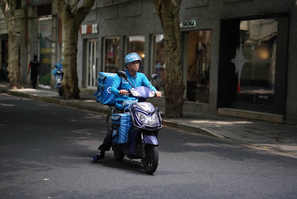 A delivery person wearing a blue uniform and helmet rides a scooter with large blue delivery boxes on a quiet street lined with trees and shops.