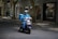 A delivery person wearing a blue uniform and helmet rides a scooter with large blue delivery boxes on a quiet street lined with trees and shops.