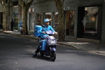 A delivery person wearing a blue uniform and helmet rides a scooter with large blue delivery boxes on a quiet street lined with trees and shops.