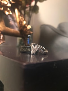 A close-up of two rings placed on a reflective surface. One ring appears to be a silver engagement ring with a large, prominent gemstone in the center, while the other has a textured band. The background features a blurred image in a decorative frame with golden accents, along with some indistinct objects.