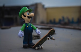 A toy figure resembling a character dressed in a green hat and blue overalls stands on a flat surface next to a skateboard. The figure appears to be looking upwards with a curious expression. The background is blurred, with muted tones giving the scene a slight overcast ambiance.