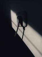 A dimly lit scene featuring a plug socket with cables attached, set against a wall. Sharp shadows create a dramatic contrast with patches of light.