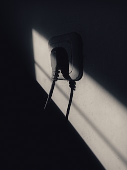 A dimly lit scene featuring a plug socket with cables attached, set against a wall. Sharp shadows create a dramatic contrast with patches of light.
