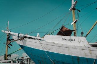 A large wooden boat with a weathered appearance is docked at a harbor, displaying the name 'GRYA IDOLA' on its white and blue hull. The ship features multiple tall masts with ropes and various rigging attached. The sky is clear with a warm turquoise hue.