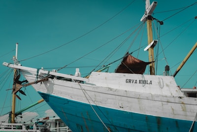 A large wooden boat with a weathered appearance is docked at a harbor, displaying the name 'GRYA IDOLA' on its white and blue hull. The ship features multiple tall masts with ropes and various rigging attached. The sky is clear with a warm turquoise hue.