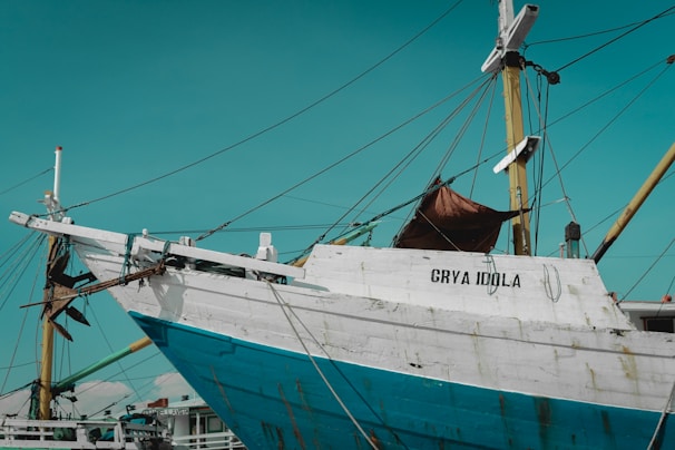 A large wooden boat with a weathered appearance is docked at a harbor, displaying the name 'GRYA IDOLA' on its white and blue hull. The ship features multiple tall masts with ropes and various rigging attached. The sky is clear with a warm turquoise hue.