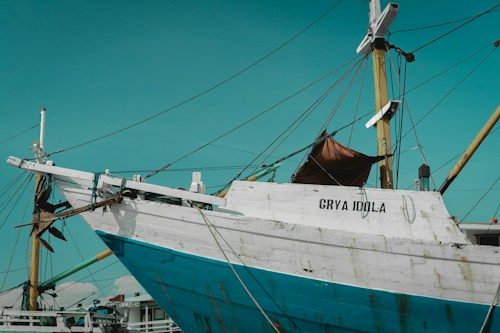 A large wooden boat with a weathered appearance is docked at a harbor, displaying the name 'GRYA IDOLA' on its white and blue hull. The ship features multiple tall masts with ropes and various rigging attached. The sky is clear with a warm turquoise hue.