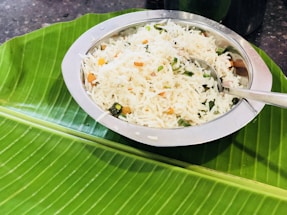 A metal bowl filled with rice mixed with vegetables such as carrots and green beans is placed atop a large banana leaf. A spoon rests in the bowl, ready for use. The setting appears to be a traditional serving style, often seen in South Asian cuisine.