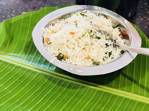 A metal bowl filled with rice mixed with vegetables such as carrots and green beans is placed atop a large banana leaf. A spoon rests in the bowl, ready for use. The setting appears to be a traditional serving style, often seen in South Asian cuisine.