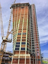 A tall building under construction with a complex scaffolding system and multiple cranes surrounding it. The structure is partially covered with orange tarps, and some areas have glass windows installed. The building appears to be in the midst of progressing towards completion.