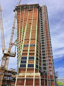 A tall building under construction with a complex scaffolding system and multiple cranes surrounding it. The structure is partially covered with orange tarps, and some areas have glass windows installed. The building appears to be in the midst of progressing towards completion.