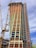 A tall building under construction with a complex scaffolding system and multiple cranes surrounding it. The structure is partially covered with orange tarps, and some areas have glass windows installed. The building appears to be in the midst of progressing towards completion.