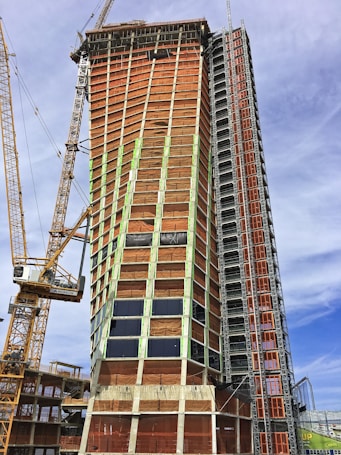 A tall building under construction with a complex scaffolding system and multiple cranes surrounding it. The structure is partially covered with orange tarps, and some areas have glass windows installed. The building appears to be in the midst of progressing towards completion.