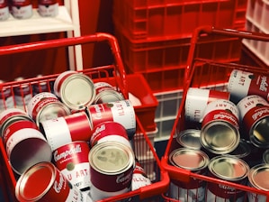 Two red shopping baskets are filled with cans resembling Campbell's soup, with stacked displays in the background. The setting appears vibrant and artistic, featuring classic pop art elements.