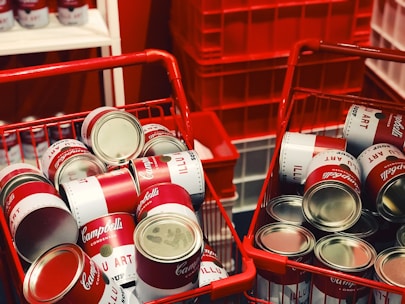 Two red shopping baskets are filled with cans resembling Campbell's soup, with stacked displays in the background. The setting appears vibrant and artistic, featuring classic pop art elements.
