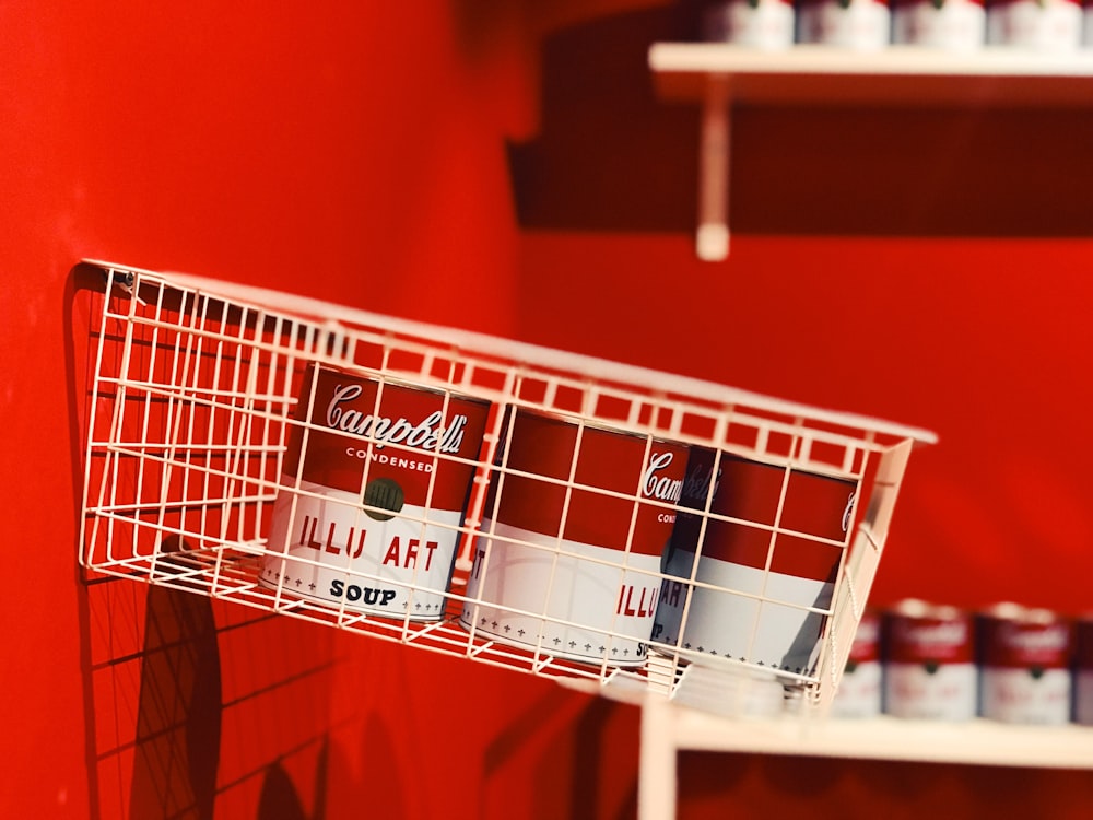 a red wall with a white wire basket holding cans of coca - cola