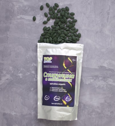 A package of spirulina tablets lies on a grey textured surface. The package is white with a colorful label displaying text and graphics. Green tablets spill out from the top of the package, scattering across the surface.