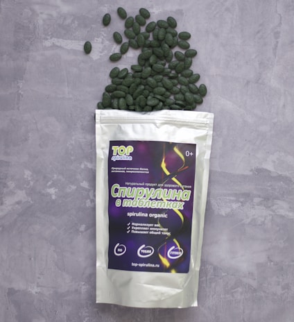 A package of spirulina tablets lies on a grey textured surface. The package is white with a colorful label displaying text and graphics. Green tablets spill out from the top of the package, scattering across the surface.