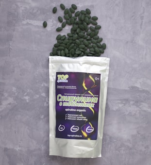 A package of spirulina tablets lies on a grey textured surface. The package is white with a colorful label displaying text and graphics. Green tablets spill out from the top of the package, scattering across the surface.