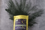 A package of spirulina powder is prominently displayed, with the bag featuring a yellow color and various text in Cyrillic. The background consists of a textured gray surface with green spirulina powder artistically spread out behind the package.