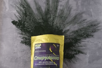 A package of spirulina powder is prominently displayed, with the bag featuring a yellow color and various text in Cyrillic. The background consists of a textured gray surface with green spirulina powder artistically spread out behind the package.