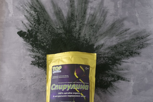 A package of spirulina powder is prominently displayed, with the bag featuring a yellow color and various text in Cyrillic. The background consists of a textured gray surface with green spirulina powder artistically spread out behind the package.