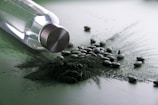 A glass bottle with a metallic cap is lying on its side, surrounded by a pile of dark green powder and scattered tablets. The setting has a reflective surface which gives a glossy appearance.