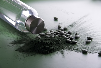 A glass bottle with a metallic cap is lying on its side, surrounded by a pile of dark green powder and scattered tablets. The setting has a reflective surface which gives a glossy appearance.