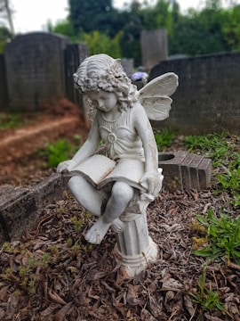 A detailed statue of a fairy-like figure with wings is seated on a pedestal, holding an open book. The figure is situated within a cemetery, surrounded by several tombstones and overgrown vegetation. The scene is marked by a mixture of brown fallen leaves, patches of green grass, and blurred gravestones in the background.