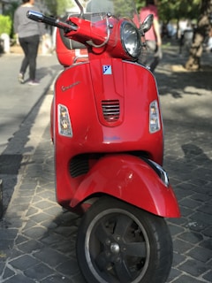 A bright red scooter is parked on a cobblestone path with sunlight casting shadows around it. The scooter has a parking sticker on its front and displays a modern and sleek design. There are people walking in the background along a shaded pedestrian path lined with trees.