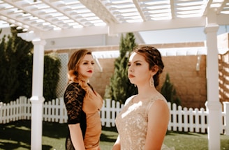 Two women dressed elegantly are standing in an outdoor setting under a white pergola. One woman is wearing a golden lace dress and the other has a copper-colored dress with black lace sleeves. A white picket fence and green trees are visible in the background, creating a serene and formal atmosphere.
