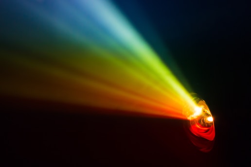 A spectrum of colored light beams spreads outward from a light source, creating an array of vibrant colors in the dark. The colors transition smoothly from red to orange, yellow, green, and blue.