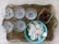 A decorative metal tray holds a set of six clear wine glasses with floral etchings. Beside them, a glass square dish contains assorted olives. A turquoise bowl filled with white, round rice crackers is also placed on the tray.
