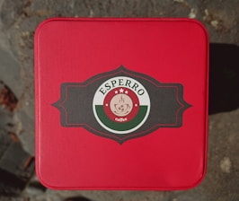 A red square tin container with rounded corners featuring a central logo. The logo consists of a black ornate frame with a circular emblem in the middle. The outer ring of the emblem is green and white while the inner circle is red containing a coffee cup with steam rising above it. The top part of the logo contains the text 'ESPERO' with three stars above it and the word 'coffee' written below the cup.