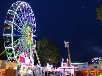 A brightly lit Ferris wheel stands prominently against a nighttime sky, adorned with vibrant, colorful lights. The scene captures a lively fairground atmosphere with various amusement rides and tents visible. A crowd of people is seen walking around, enjoying the festivities.