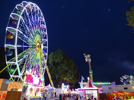 A brightly lit Ferris wheel stands prominently against a nighttime sky, adorned with vibrant, colorful lights. The scene captures a lively fairground atmosphere with various amusement rides and tents visible. A crowd of people is seen walking around, enjoying the festivities.