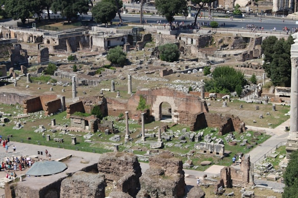 Aerial view of ancient ruins featuring a mix of standing columns and fragmented structures. The area is surrounded by vegetation, and pathways are visible among the ruins. People can be seen exploring the site, indicating a popular tourist destination. The scene is set with trees and modern roads in the background.
