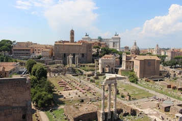 An ancient urban landscape featuring well-preserved ruins and classical architecture, including towering columns, arches, and various historical buildings. The background showcases a blend of ancient and more modern edifices under a clear blue sky.