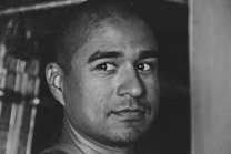 A black and white portrait of a person with a shaved head looking directly at the camera. The background is blurred, and the focus is on the individual's face, which has a neutral yet slightly serious expression. The lighting casts soft shadows, highlighting facial features.