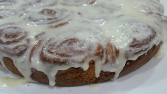 A close-up of a baked cinnamon roll cake topped with a thick layer of creamy icing that is slightly dripping down the sides. The swirled pattern of the cinnamon rolls is visible through the glaze.