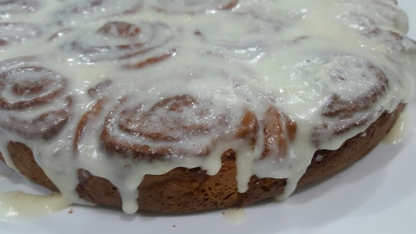 A close-up of a baked cinnamon roll cake topped with a thick layer of creamy icing that is slightly dripping down the sides. The swirled pattern of the cinnamon rolls is visible through the glaze.
