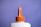 A white plastic bottle with an orange twist cap and a pointed nozzle, set against a solid purple background. The image focuses on the top portion of the bottle, highlighting the cap's ridged texture and the nozzle's cone shape.