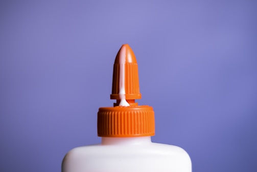A white plastic bottle with an orange twist cap and a pointed nozzle, set against a solid purple background. The image focuses on the top portion of the bottle, highlighting the cap's ridged texture and the nozzle's cone shape.