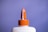 A white plastic bottle with an orange twist cap and a pointed nozzle, set against a solid purple background. The image focuses on the top portion of the bottle, highlighting the cap's ridged texture and the nozzle's cone shape.
