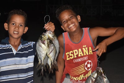 Two boys are standing together, proudly holding several fish. The boy on the left is wearing a blue-striped shirt, while the boy on the right is wearing a red tank top with the word 'España' written on it. Both are smiling broadly, suggesting they had a successful fishing trip or activity and are happy with their catch. It is nighttime or in a dimly lit area.