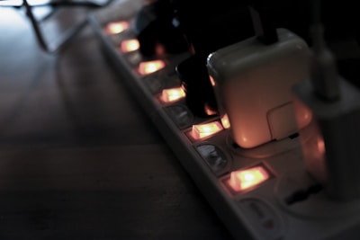 A power strip with several electronic devices plugged in, illuminated by small orange indicator lights. The scene is dimly lit, casting a shadowy atmosphere over a wooden surface.