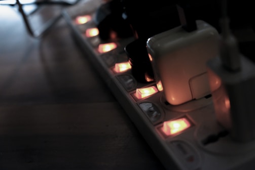 A power strip with several electronic devices plugged in, illuminated by small orange indicator lights. The scene is dimly lit, casting a shadowy atmosphere over a wooden surface.