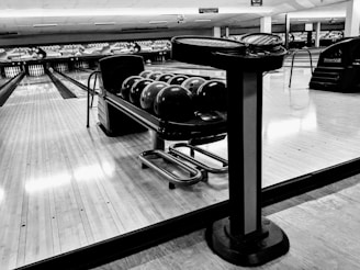 A row of bowling balls set on a rack near lanes in a bowling alley with a low ceiling. The polished wooden lane floor reflects overhead lights. A stand with a scorekeeping screen is positioned nearby. The setting is empty, conveying a sense of stillness.